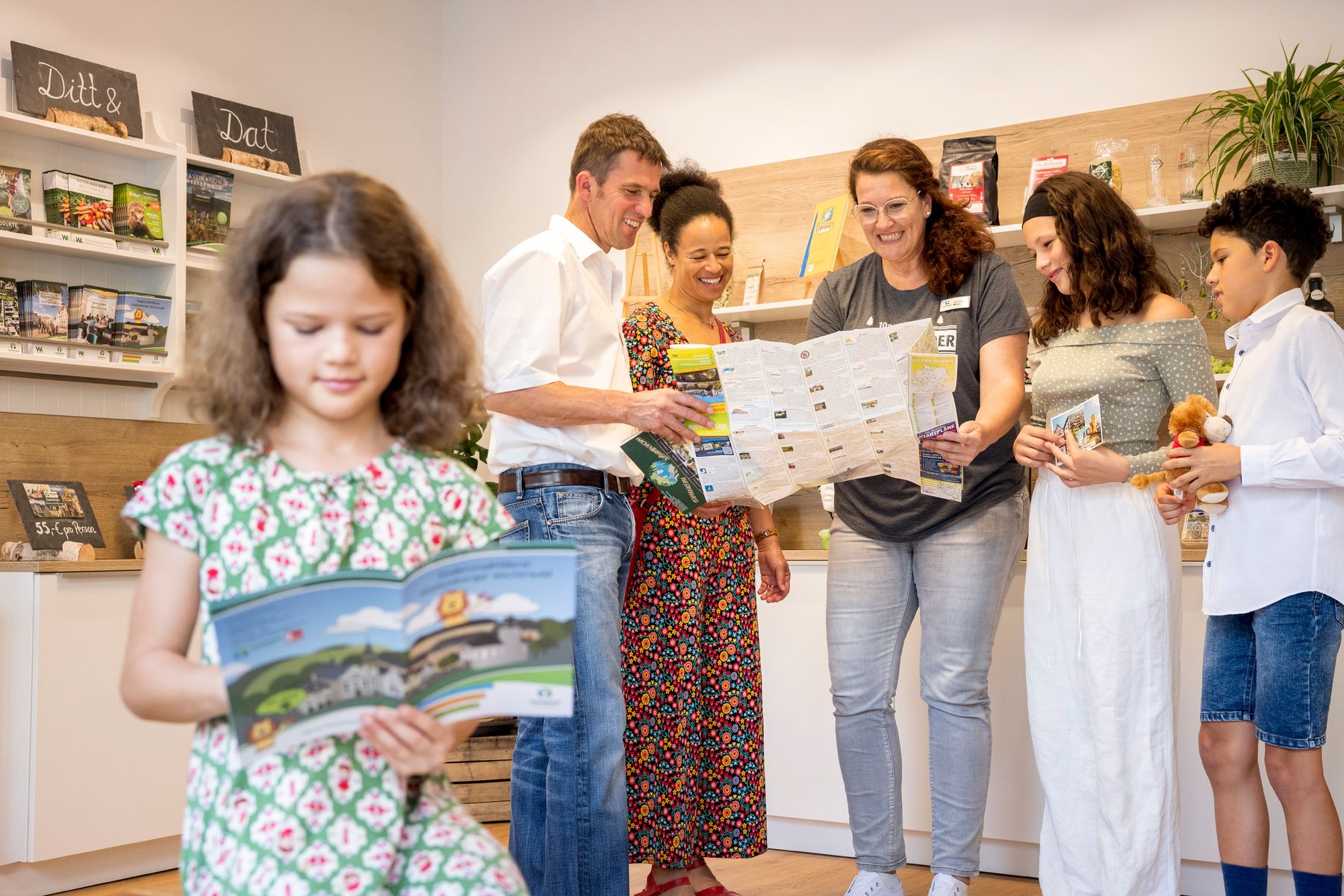 Eine Gruppe von Erwachsenen und Kindern steht in einem Geschäft, lächelt und sieht sich Broschüren und Karten an. Im Hintergrund sind Regale mit Produkten und Informationsmaterial zu sehen. Ein Mädchen hält eine Broschüre im Vordergrund.